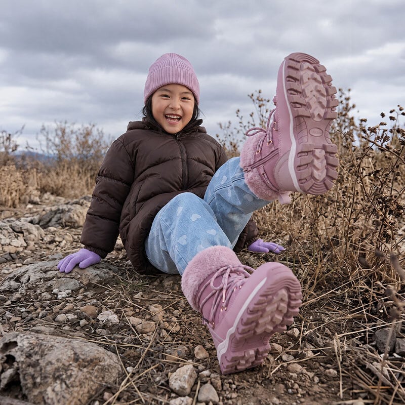 En tjej sitter på marken med varm jacka, lila mössa och vantar och varmfodrade boots